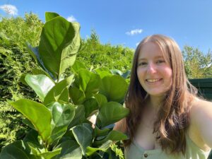 Picture of Brianna and a green Fiddle Leaf Fig plant with big leaves outside on a sunny day. Fiddle Leaf Fig Care to help keep your plant alive.